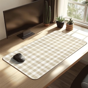 Puede incluir: Alfombrilla de escritorio con estampado de cuadros beige y blanco, con un ratón de ordenador negro en el lado izquierdo. La alfombrilla rectangular está sobre un escritorio de madera. Un monitor de ordenador y plantas en el fondo.