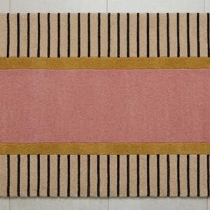 Op de afbeelding: Een ovaalvormig loperkleed met een effen roze middenstuk, een mosterdgele rand en een buitenrand met afwisselende beige en zwarte verticale strepen. Het kleed heeft een getufte textuur en een symmetrisch ontwerp met afgeronde uiteinden.