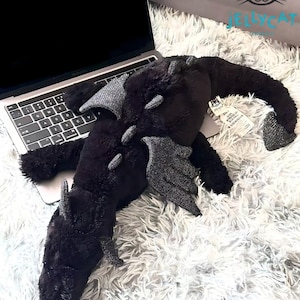 May include: A black plush dragon toy with sparkly grey detailing on its wings, horns, and spine. The dragon is positioned on a white, fluffy surface next to a laptop. The toy has a tag attached. The background includes a bag with the text "AMUSEABLES" and "JELLYCAT".