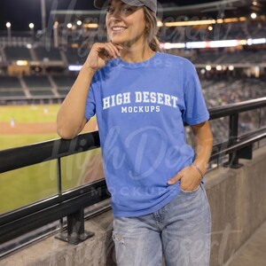 Peut inclure: Un t-shirt bleu &agrave; manches courtes avec le texte blanc HIGH DESERT MOCKUPS imprim&eacute; sur la poitrine, associ&eacute; &agrave; une casquette de baseball grise et un jean bleu clair d&eacute;lav&eacute;, port&eacute; dans un stade avec un terrain flou en arri&egrave;re-plan.