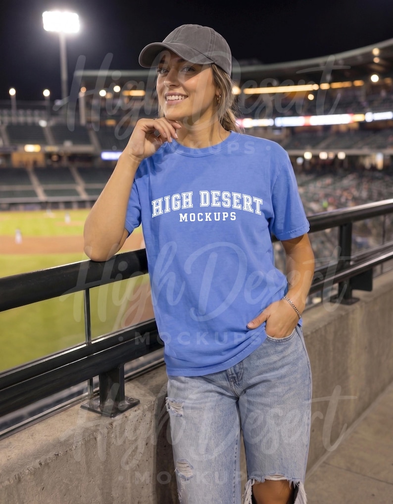 Peut inclure: Un t-shirt bleu &agrave; manches courtes avec le texte blanc HIGH DESERT MOCKUPS imprim&eacute; sur la poitrine, associ&eacute; &agrave; une casquette de baseball grise et un jean bleu clair d&eacute;lav&eacute;, port&eacute; dans un stade avec un terrain flou en arri&egrave;re-plan.