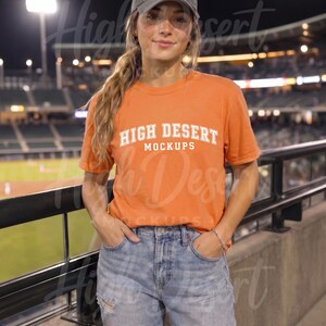 Peut inclure: Une casquette de baseball gris anthracite et un t-shirt orange &agrave; manches courtes avec le texte blanc HIGH DESERT MOCKUPS dans une police universitaire, associ&eacute;s &agrave; un jean bleu clair avec des d&eacute;tails us&eacute;s sur le devant des jambes.