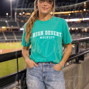 Peut inclure: Un t-shirt &agrave; manches courtes vert vif avec le texte blanc HIGH DESERT MOCKUPS imprim&eacute; sur la poitrine, associ&eacute; &agrave; un jean bleu clair d&eacute;lav&eacute; et une casquette de baseball gris fonc&eacute;, sur un fond de stade flou.