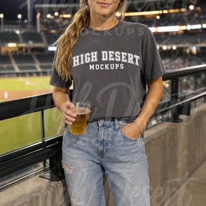 Peut inclure: Un t-shirt gris anthracite &agrave; manches courtes avec le texte blanc HIGH DESERT MOCKUPS imprim&eacute; sur la poitrine, associ&eacute; &agrave; un jean bleu clair d&eacute;lav&eacute; et une casquette de baseball gris anthracite assortie, port&eacute; dans un stade.