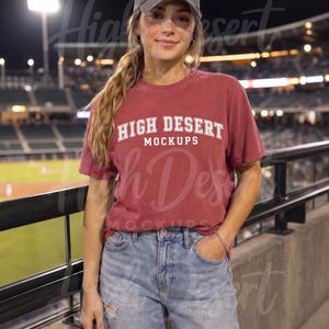 Peut inclure: Un t-shirt &agrave; manches courtes rouge fonc&eacute; avec le texte blanc HIGH DESERT MOCKUPS imprim&eacute; sur la poitrine, associ&eacute; &agrave; un jean bleu clair d&eacute;lav&eacute; et une casquette de baseball gris fonc&eacute;, port&eacute; dans un stade la nuit.