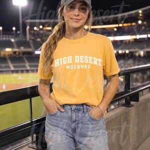 Peut inclure: Un t-shirt &agrave; manches courtes jaune moutarde avec le texte blanc HIGH DESERT MOCKUPS imprim&eacute; sur la poitrine. Le haut est associ&eacute; &agrave; une casquette de baseball gris anthracite et &agrave; un jean en denim bleu clair us&eacute; avec des d&eacute;chirures aux genoux.