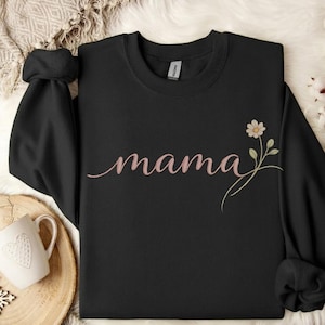 Puede incluir: Sudadera negra con la palabra "mama" en rosa en cursiva. Una pequeña margarita con hojas verdes adorna la prenda. Las mangas están dobladas. La sudadera está doblada.