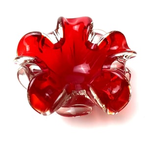 Può includere: Un posacenere in vetro rosso vibrante con un design unico a forma di fiore. Il posacenere presenta sei petali arrotondati e una base in vetro trasparente, creando un contrasto sorprendente. La finitura lucida esalta il colore rosso intenso.