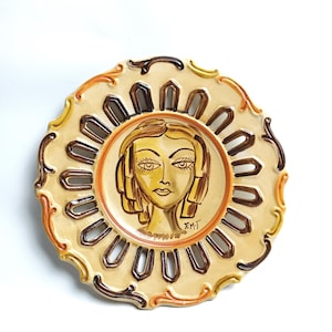 Può includere: Un piatto decorativo rotondo in ceramica con un'illustrazione centrale dipinta a mano di un volto con capelli stilizzati. Il piatto ha una base color crema con accenti marroni e arancioni, un bordo smerlato e fessure ritagliate. Firmato F.M.T.