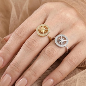 May include: Two rings featuring compass designs. One is gold-coloured with a compass and a halo of small stones. The other is silver-coloured with a compass and a halo of small stones. The rings are on a hand.