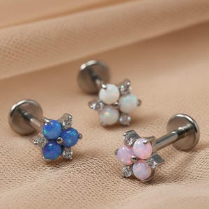 Könnte beinhalten: Drei blütenförmige Ohr- oder Lippenstecker. Einer hat blaue Opalsteine, ein weiterer weiße und der dritte rosa. Jeder Stecker hat einen silbernen Stift und ist mit kleinen, klaren Kristallen verziert. Der Hintergrund ist ein weicher, neutraler Stoff.