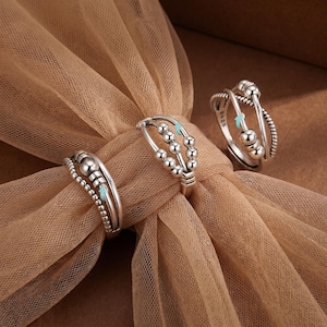 May include: Three silver fidget rings with beaded and textured designs. Each ring features small, round beads and a light blue accent. The rings are displayed on a tan fabric, showcasing their intricate details and metallic shine.