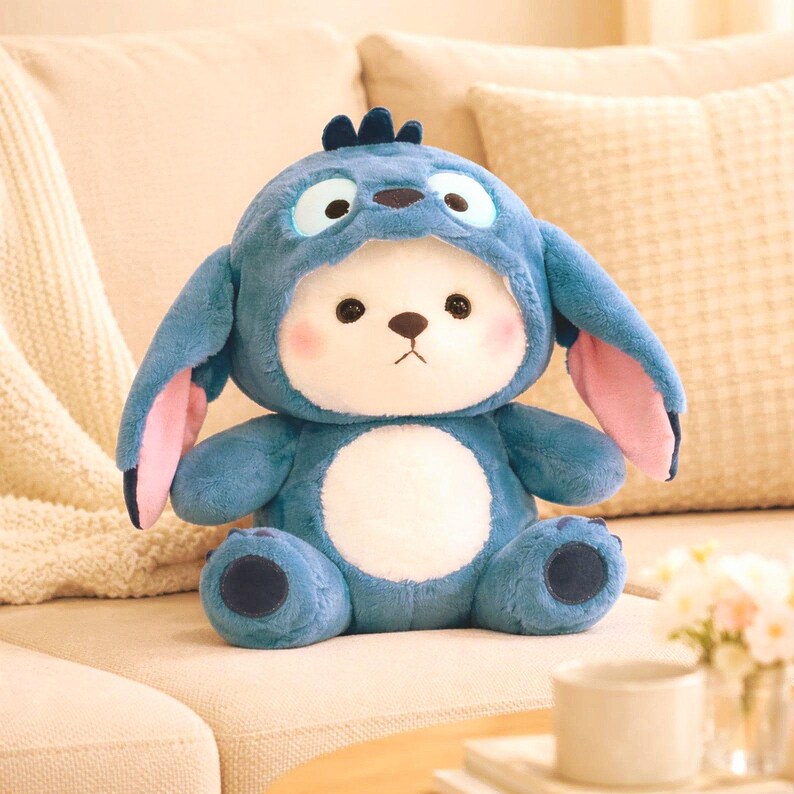 May include: A plush toy in the shape of a bear wearing a blue costume with large ears and a tuft of hair. The bear has a white face with pink cheeks, black eyes, and a brown nose. The toy is sitting on a beige couch.