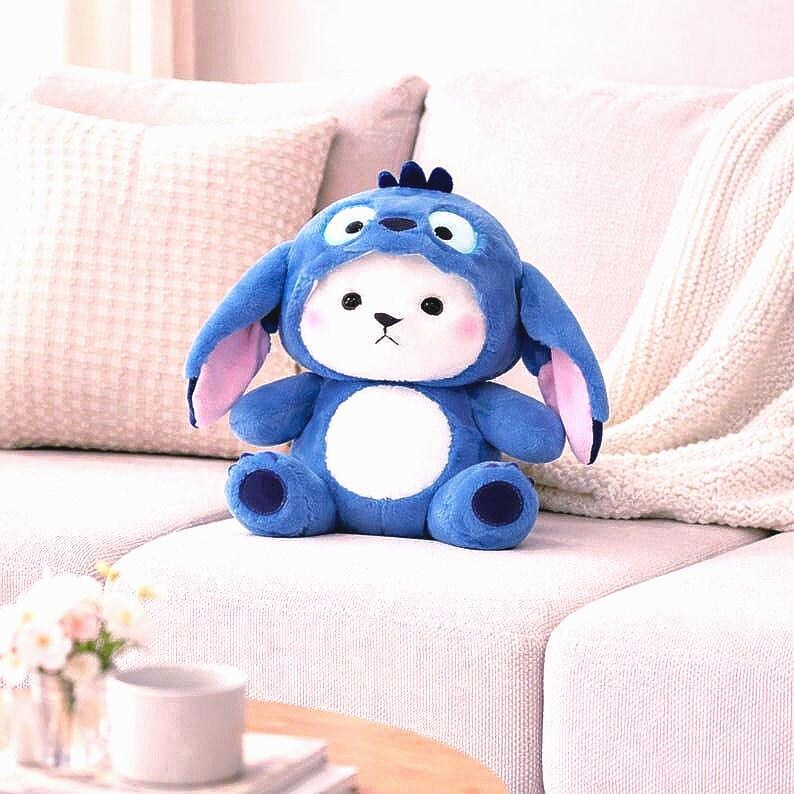 May include: A plush blue stuffed animal in the shape of a character with large ears and a white belly. The toy has a blue hood with large eyes and a small tuft of hair. The ears are lined with pink fabric. The toy is sitting on a white couch.