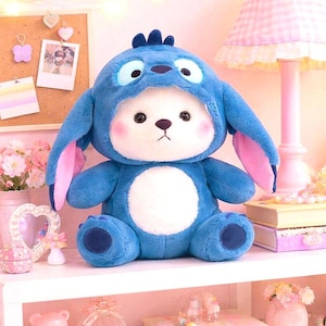 May include: A plush blue stuffed animal with a white face and a blue hood resembling a cartoon character. The toy has large, round eyes, pink cheeks, and large ears with pink inner ears. It is sitting on a white shelf with other decorative items.