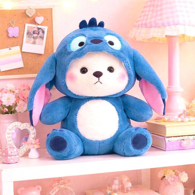 May include: A plush blue stuffed animal with a white face and a blue hood resembling a cartoon character. The toy has large, round eyes, pink cheeks, and large ears with pink inner ears. It is sitting on a white shelf with other decorative items.