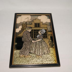 May include: A framed illustration of a couple in traditional Dutch clothing standing in front of a house with a stork on the roof. The illustration is done in gold and silver metallic paint.