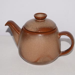 May include: A brown ceramic teapot with a lid and handle. The teapot has a rounded body and a spout. The lid has a small knob on top. The teapot is a warm brown color with darker brown accents.