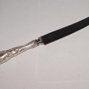 May include: A silver butter knife with an ornate handle and a dark blade. The handle features a detailed floral design. The knife is set against a plain, light-colored background, highlighting its elegant design. This utensil is suitable for serving butter or other spreads.