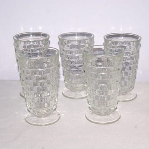 May include: Set of five clear, textured drinking glasses. Each glass has a footed base and a cylindrical body with a grid-like pattern. The glasses are arranged on a white surface.