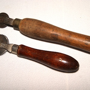 May include: Two vintage sewing tracing wheels with wooden handles. One handle is a lighter brown, the other is a darker brown. The wheels are metal and have a serrated edge. The tools are likely used for transferring pattern markings.