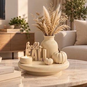 Könnte beinhalten: Ein cremefarbenes, rundes Tablett mit einer gerippten Vase voller getrocknetem Pampasgras, zwei kleinen cremefarbenen Kürbissen, einer brennenden Kerze in einem gerippten Halter und einem Holzschild mit dem Text hello FALL in Blockschrift.