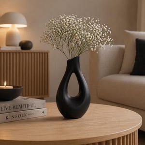 Könnte beinhalten: Eine schwarze, moderne Vase mit einzigartigem Ausschnitt-Design hält weiße Blumen. Eine brennende Kerze steht in einer schwarzen Schale auf zwei Büchern. Die Szene spielt sich auf einem runden, hellen Holztisch ab.
