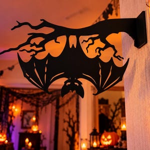 Pode incluir: Uma decoração de parede em metal preto com a silhueta de um morcego pendurado de cabeça para baixo num galho de árvore nu. O design é plano e recortado, mostrando o morcego com as asas abertas e orelhas pontiagudas fixado na parede.