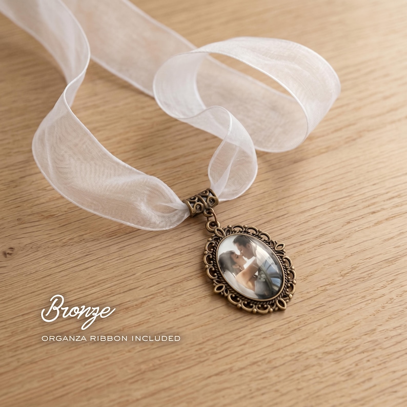 May include: An oval bronze pendant featuring an ornate filigree frame and a central photograph of a couple, suspended from a sheer white organza ribbon. The text Bronze and ORGANZA RIBBON INCLUDED is printed on the image in white lettering.