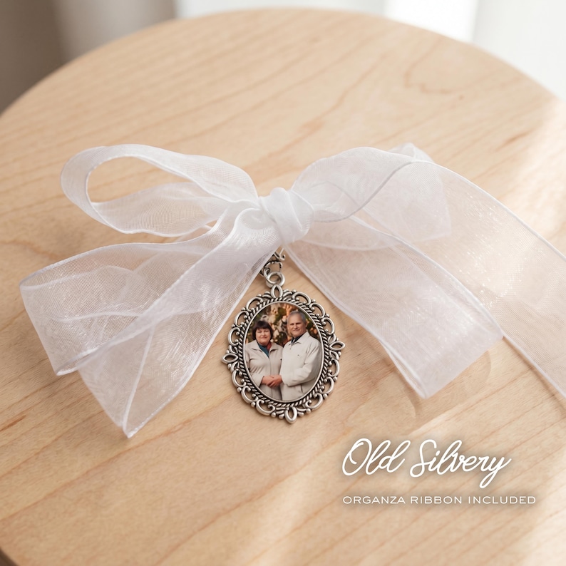 May include: An oval-shaped silver-toned metal pendant featuring an intricate filigree border and a photograph of two people inside. The pendant is attached to a large, sheer white organza ribbon tied into a decorative bow with long flowing ends.