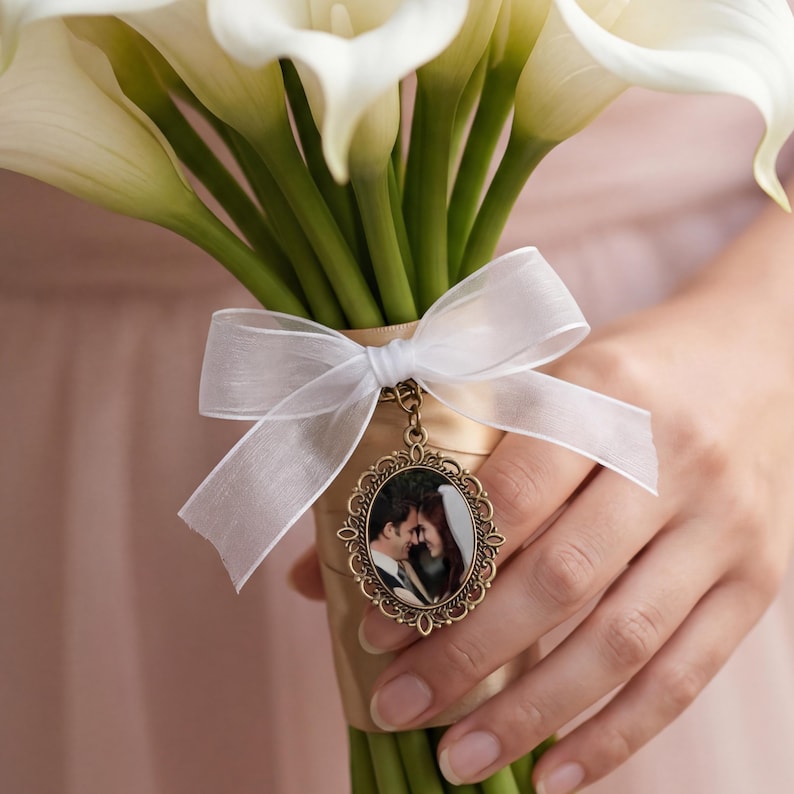 May include: A bouquet of white calla lilies wrapped in tan ribbon with a white sheer organza bow. An oval bronze-toned metal locket featuring a photograph of two people is attached to the ribbon, hanging from a small metal ring below the bow.