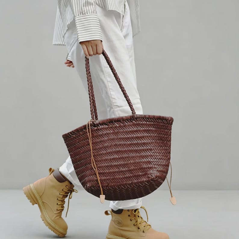 Peut inclure: Un sac fourre-tout marron tress&eacute; avec des anses tress&eacute;es. Le sac a une forme arrondie et est tenu par une personne portant un pantalon blanc et des bottes beiges. La texture du sac est visible, montrant le motif de tissage complexe.