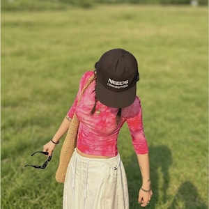 Puede incluir: Una mujer con una gorra marr&oacute;n con el texto "NEEDS Connecting People", una camiseta rosa de manga larga con te&ntilde;ido anudado y una falda blanca. Lleva un bolso tejido y sostiene gafas de sol. El fondo es un campo verde.