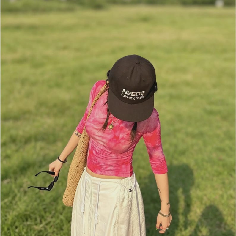 Puede incluir: Una mujer con una gorra marr&oacute;n con el texto "NEEDS Connecting People", una camiseta rosa de manga larga con te&ntilde;ido anudado y una falda blanca. Lleva un bolso tejido y sostiene gafas de sol. El fondo es un campo verde.