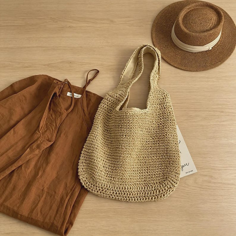 Puede incluir: Un bolso tote de paja tejida con interior beige se encuentra junto a un sombrero para el sol marr&oacute;n con una banda blanca. Un vestido color &oacute;xido est&aacute; doblado sobre una superficie de madera. La bolsa est&aacute; abierta y vac&iacute;a.