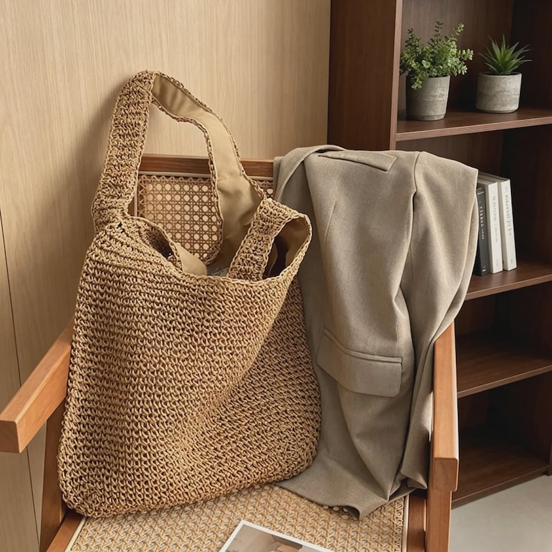 Puede incluir: Un bolso tote de paja tejida con interior beige est&aacute; sobre una silla de madera. Una blazer color beige est&aacute; sobre el respaldo de la silla. El bolso tiene un dise&ntilde;o grande y abierto y correas gruesas.