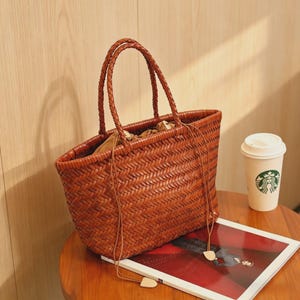 Peut inclure: Un sac fourre-tout en cuir tress&eacute; marron avec des poign&eacute;es tress&eacute;es. Le sac a une forme rectangulaire et une fermeture &agrave; cordon. Une tasse en papier blanche avec un logo vert est pos&eacute;e sur une table.