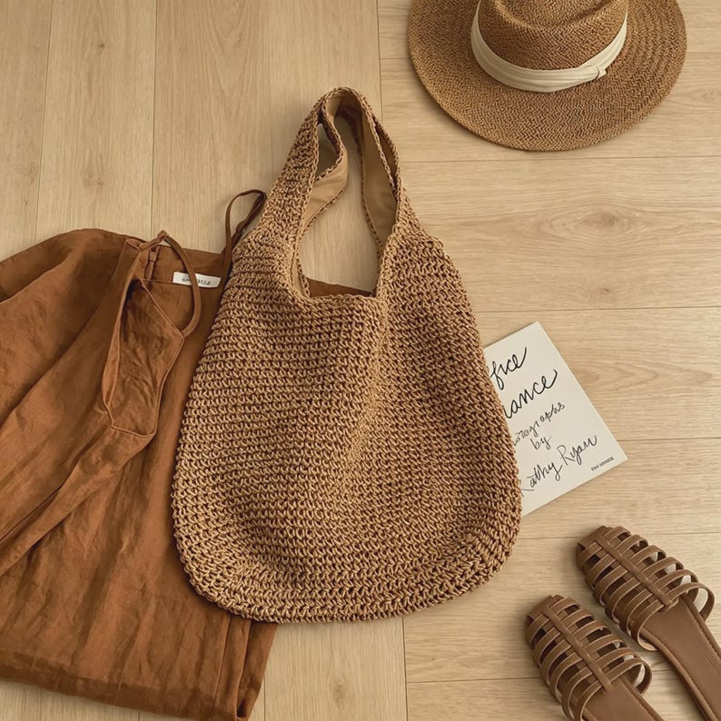 Puede incluir: Un bolso tote tejido marr&oacute;n con una correa ancha para el hombro se encuentra junto a una camisa de lino marr&oacute;n, un sombrero de paja con una cinta blanca y sandalias marrones. Tambi&eacute;n es visible una peque&ntilde;a tarjeta con texto manuscrito.