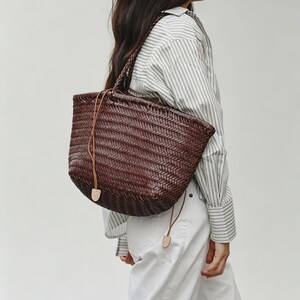 Peut inclure: Un sac fourre-tout en cuir tress&eacute; marron fonc&eacute; avec une poign&eacute;e tress&eacute;e et une fermeture &agrave; cordon. Le sac a une forme arrondie et se porte &agrave; l'&eacute;paule. Le sac est en cuir marron fonc&eacute;.