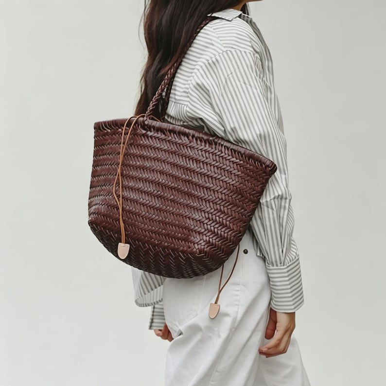 Peut inclure: Un sac fourre-tout en cuir tress&eacute; marron fonc&eacute; avec une poign&eacute;e tress&eacute;e et une fermeture &agrave; cordon. Le sac a une forme arrondie et se porte &agrave; l'&eacute;paule. Le sac est en cuir marron fonc&eacute;.