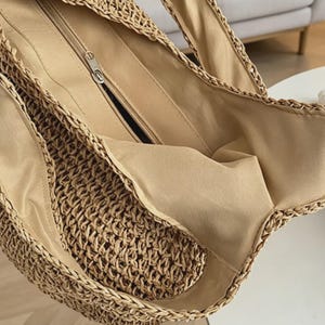 Puede incluir: Vista de cerca de un bolso tote de paja tejida. El bolso tiene un forro interior beige y un bolsillo con cremallera. Las asas est&aacute;n hechas del mismo material tejido que el bolso. El bolso est&aacute; dise&ntilde;ado para transportar objetos.