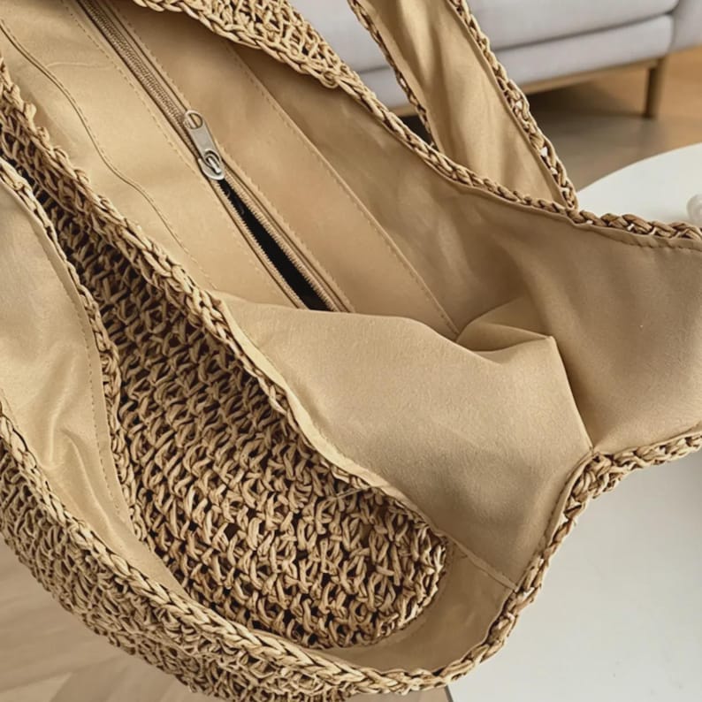Puede incluir: Vista de cerca de un bolso tote de paja tejida. El bolso tiene un forro interior beige y un bolsillo con cremallera. Las asas est&aacute;n hechas del mismo material tejido que el bolso. El bolso est&aacute; dise&ntilde;ado para transportar objetos.