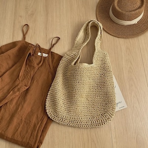 Puede incluir: Un bolso tote tejido beige con asas cortas est&aacute; junto a un vestido de verano marr&oacute;n y un sombrero de paja con una cinta blanca. Los art&iacute;culos est&aacute;n colocados sobre una superficie de madera clara.