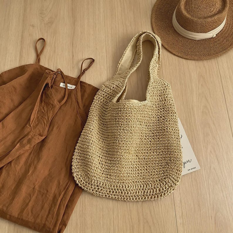 Puede incluir: Un bolso tote tejido beige con asas cortas est&aacute; junto a un vestido de verano marr&oacute;n y un sombrero de paja con una cinta blanca. Los art&iacute;culos est&aacute;n colocados sobre una superficie de madera clara.