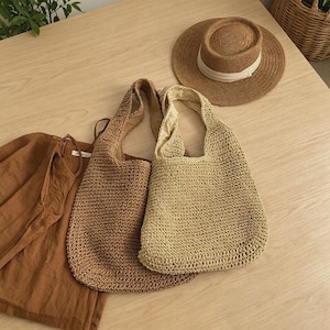 Puede incluir: Dos bolsos tote tejidos en marr&oacute;n claro y beige, junto con un sombrero de paja con una banda blanca. Tambi&eacute;n se ve una camisa de lino marr&oacute;n. Los art&iacute;culos est&aacute;n dispuestos sobre una superficie de madera clara, sugiriendo un tema de verano o playa.