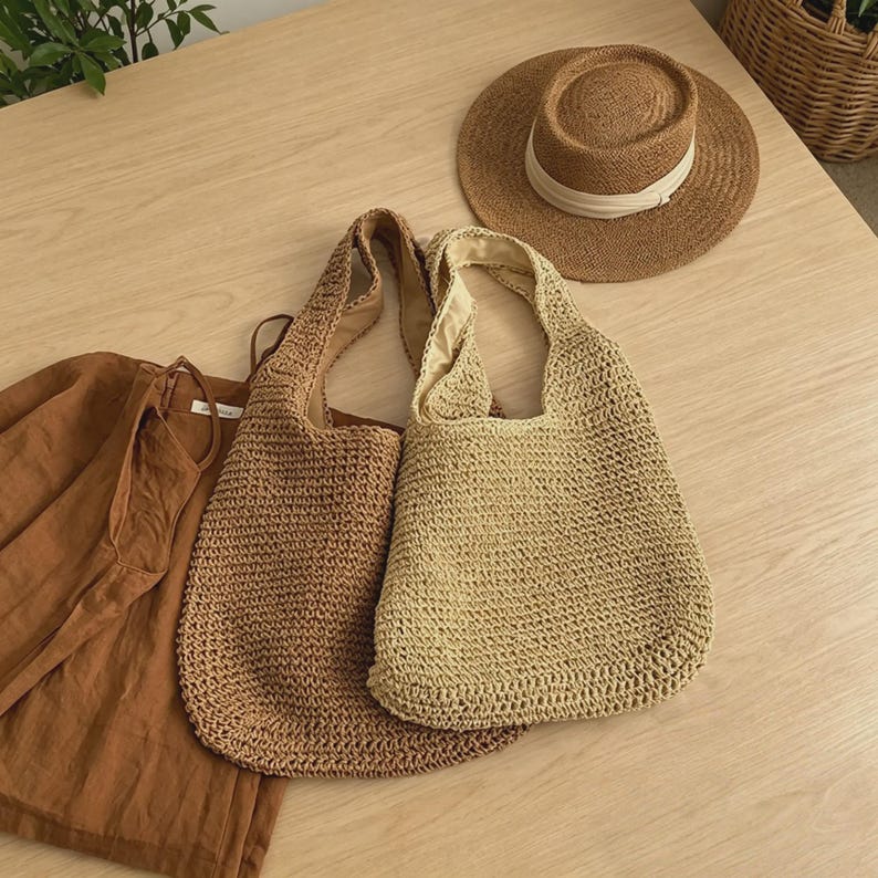Puede incluir: Dos bolsos tote tejidos en marr&oacute;n claro y beige, junto con un sombrero de paja con una banda blanca. Tambi&eacute;n se ve una camisa de lino marr&oacute;n. Los art&iacute;culos est&aacute;n dispuestos sobre una superficie de madera clara, sugiriendo un tema de verano o playa.