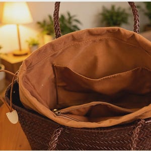 Peut inclure: Un sac fourre-tout marron tress&eacute; avec des poign&eacute;es tress&eacute;es. L'int&eacute;rieur est doubl&eacute; d'un tissu marron avec des poches et un compartiment zipp&eacute;. Le sac est ouvert, r&eacute;v&eacute;lant l'int&eacute;rieur. Le sac est con&ccedil;u pour transporter des objets.