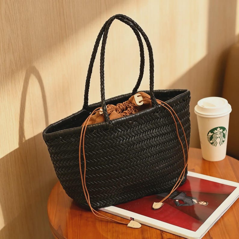 Peut inclure: Sac fourre-tout noir tiss&eacute; avec des poign&eacute;es tress&eacute;es et une fermeture &agrave; cordon. Le sac a de longues et fines lani&egrave;res marron en simili cuir avec des accents en bois. Une tasse &agrave; caf&eacute; et un magazine sont &agrave; c&ocirc;t&eacute; du sac sur une table en bois.