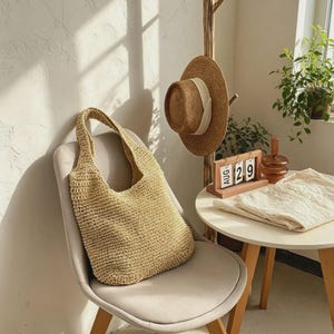 Puede incluir: Un bolso tote de paja tejida de color beige claro est&aacute; sobre una silla gris. Un sombrero de paja con una cinta blanca cuelga de un perchero de madera. Un calendario de madera muestra la fecha 29 de agosto. Una prenda de punto blanca est&aacute; sobre una mesa redonda.