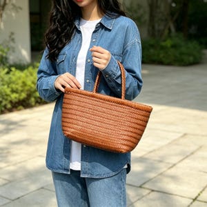 Puede incluir: Un bolso tote trenzado de color marrón de tamaño mediano, con un patrón de espiga diagonal ajustado en todo el cuerpo y dos asas trenzadas a juego. El bolso tiene una base rectangular estructurada y una parte superior abierta, hecho de material liso.